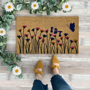 May include: A rectangular doormat with a floral design and the letters "FDT" in blue. The flowers are in shades of pink, orange, and blue. A blue butterfly is on the right side. The doormat is on a wooden floor.