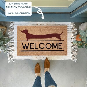 May include: A brown coir doormat with a welcome message and a red dachshund silhouette. The doormat is on a concrete floor with a pair of brown leather shoes and blue jeans.