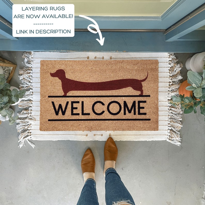 May include: A brown coir doormat with a welcome message and a red dachshund silhouette. The doormat is on a concrete floor with a pair of brown leather shoes and blue jeans.