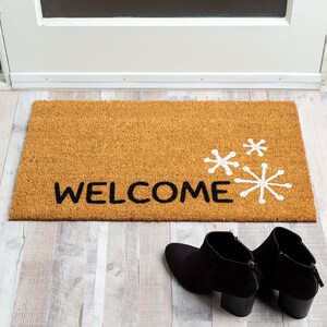 May include: A brown coir doormat with the word "WELCOME" in black lettering and white snowflake designs.