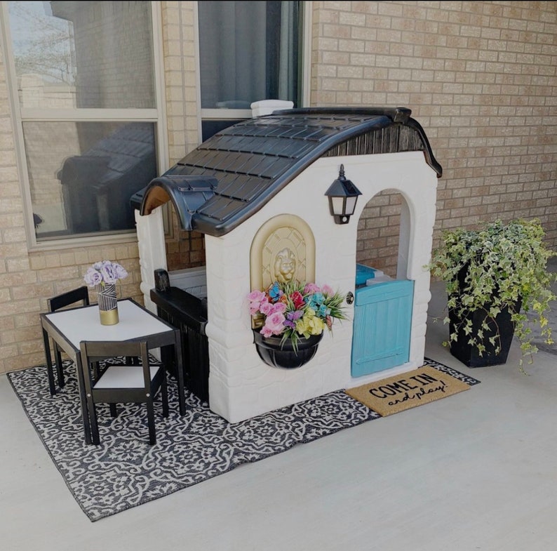 May include: A white and black playhouse with a blue door and a black and white rug. The playhouse has a black roof and a decorative fountain on the front. The playhouse has a sign that says "Come in and play".