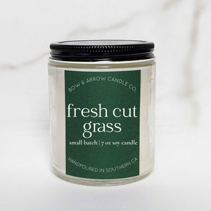 May include: A clear glass candle jar with a black lid. The label is green and says "BOW & ARROW CANDLE CO. fresh cut grass small batch | 7 oz soy candle HANDPOURED IN SOUTHERN CA"