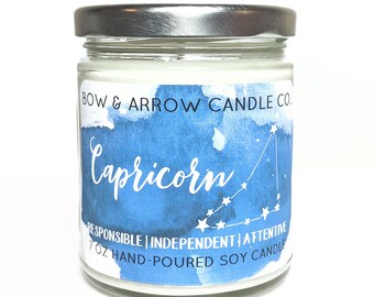 May include: A 7 ounce soy candle in a clear glass jar with a silver lid. The label is blue with white watercolor-style clouds and stars. The label says "BOW & ARROW CANDLE CO." and "Capricorn" in a white cursive font. Below the name is a constellation of stars and the words "RESPONSIBLE | INDEPENDENT | ATTENTIVE" in white. The bottom of the label says "7 OZ HAND-POURED SOY CANDLE".