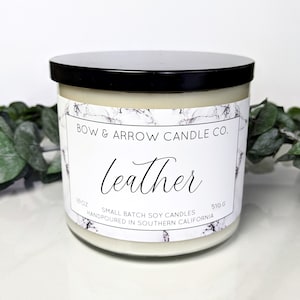 May include: A white candle jar with a black lid and a marble-patterned label. The label reads "BOW & ARROW CANDLE CO." and "leather". The candle is 18 oz and 510 g. It is a small batch soy candle handpoured in Southern California.