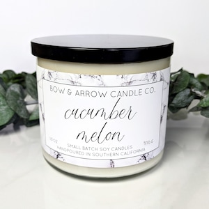 Puede incluir: Un tarro de vela blanco con una tapa negra. La etiqueta es blanca con un patrón de mármol y texto negro que dice "BOW & ARROW CANDLE CO. cucumber melon SMALL BATCH SOY CANDLES HANDPOURED IN SOUTHERN CALIFORNIA 18 oz 510 g".