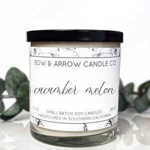 Puede incluir: Un tarro de vela blanco con una tapa negra. La etiqueta dice "BOW & ARROW CANDLE CO." y "cucumber melon". La etiqueta también dice "10 OZ SMALL BATCH SOY CANDLES HANDPOURED IN SOUTHERN CALIFORNIA 283 G"