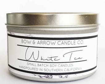 May include: A silver candle tin with a white label that reads "BOW & ARROW CANDLE CO. White Tea SMALL BATCH SOY CANDLES HANDPOURED IN SOUTHERN CALIFORNIA".