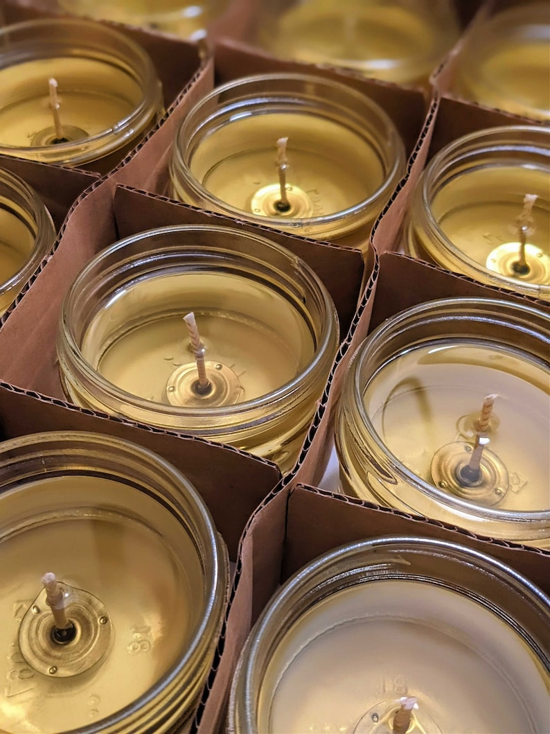 May include: Close-up of multiple glass jars filled with a golden yellow wax, each with a single wick centered in the jar. The jars are arranged in rows on a brown cardboard surface.