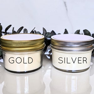 May include: Two small glass candle jars with gold and silver lids. The candle in the jar with the gold lid is labeled "GOLD" and the candle in the jar with the silver lid is labeled "SILVER".