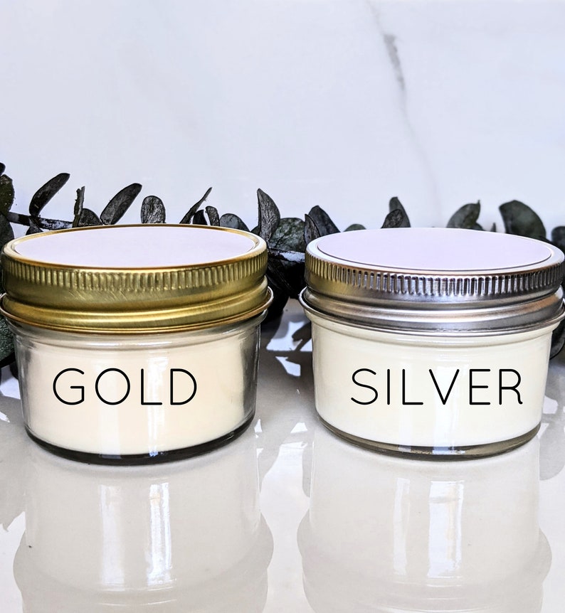 May include: Two small glass candle jars with gold and silver lids. The candle in the jar with the gold lid is labeled "GOLD" and the candle in the jar with the silver lid is labeled "SILVER".