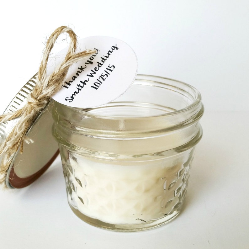 May include: A small glass jar candle with a white lid and a twine bow. A white tag with black text reads "Thank you! Smith Wedding 10/25/15".