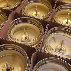 May include: Close-up of multiple glass jars filled with a golden yellow wax, each with a single wick centered in the jar. The jars are arranged in rows on a brown cardboard surface.