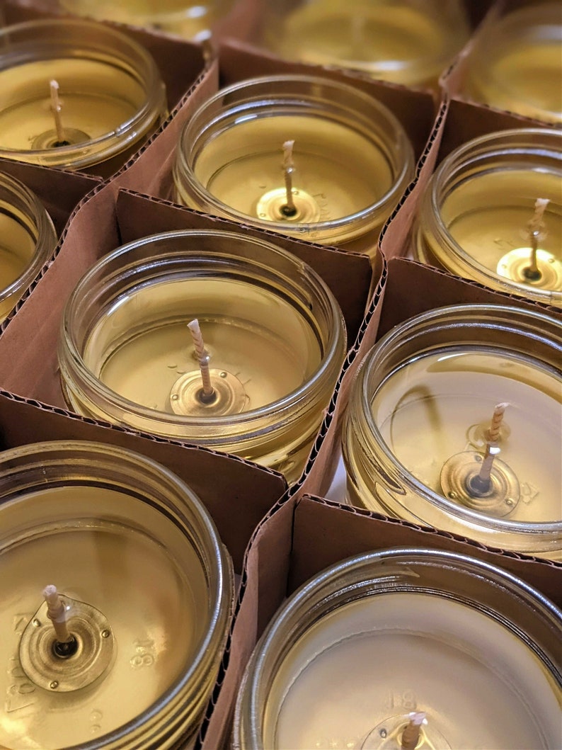 May include: Close-up of multiple glass jars filled with a golden yellow wax, each with a single wick centered in the jar. The jars are arranged in rows on a brown cardboard surface.