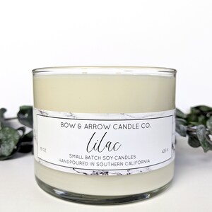 May include: A white candle in a clear glass jar with a white label that reads "BOW & ARROW CANDLE CO. lilac SMALL BATCH SOY CANDLES HANDPOURED IN SOUTHERN CALIFORNIA" and has a marble pattern. The candle is 5 oz or 425 g.