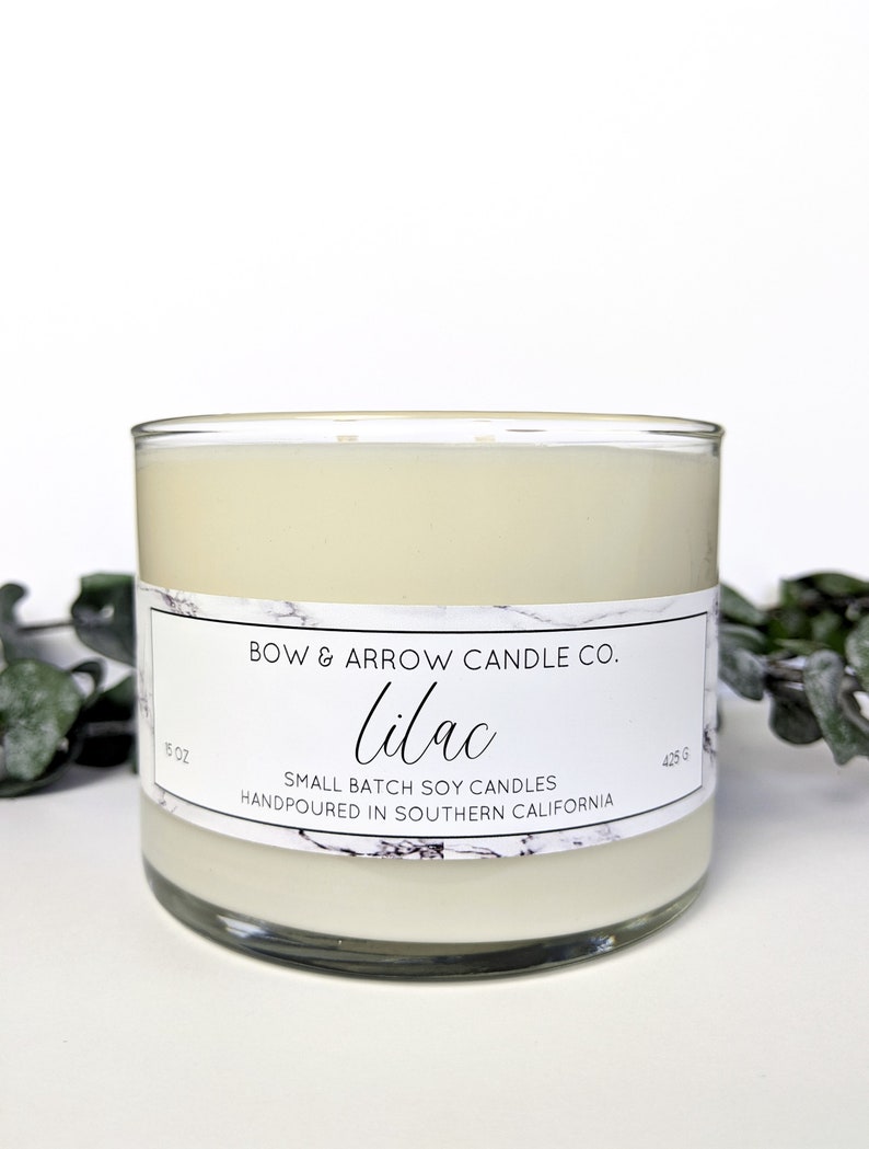 May include: A white candle in a clear glass jar with a white label that reads "BOW & ARROW CANDLE CO. lilac SMALL BATCH SOY CANDLES HANDPOURED IN SOUTHERN CALIFORNIA" and has a marble pattern. The candle is 5 oz or 425 g.