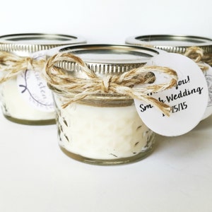 May include: Three small glass jars containing white candles. The jars have silver lids and are tied with twine and a bow. A white round tag is attached to one jar with the text "Thank you! Smith Wedding 12/15/15".