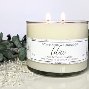 May include: A clear glass candle jar with a white label that reads "BOW & ARROW CANDLE CO. Lilac SMALL BATCH SOY CANDLES HANDPOURED IN SOUTHERN CALIFORNIA". The jar is 15 oz and 425 g. The candle is lit with two wicks.