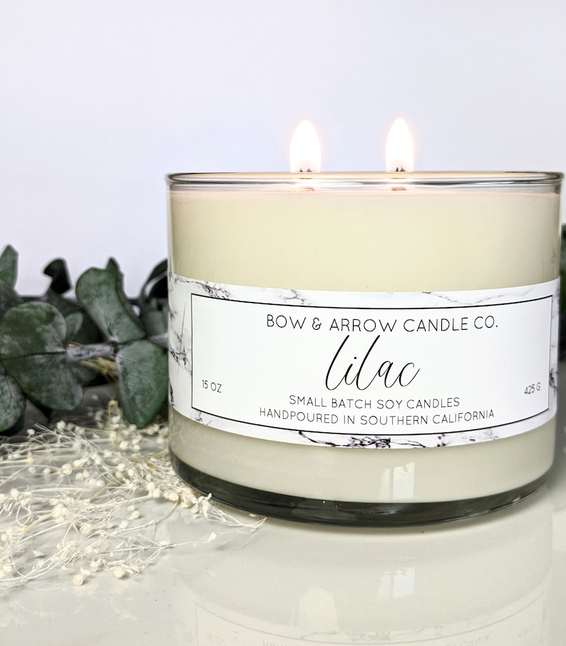May include: A clear glass candle jar with a white label that reads "BOW & ARROW CANDLE CO. Lilac SMALL BATCH SOY CANDLES HANDPOURED IN SOUTHERN CALIFORNIA". The jar is 15 oz and 425 g. The candle is lit with two wicks.