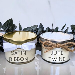 May include: Two small glass jars with white candles inside. The jars have gold and silver lids. The jar on the left has a white satin ribbon tied around the lid. The jar on the right has a brown jute twine tied around the lid. The jars are labeled "SATIN RIBBON" and "JUTE TWINE".