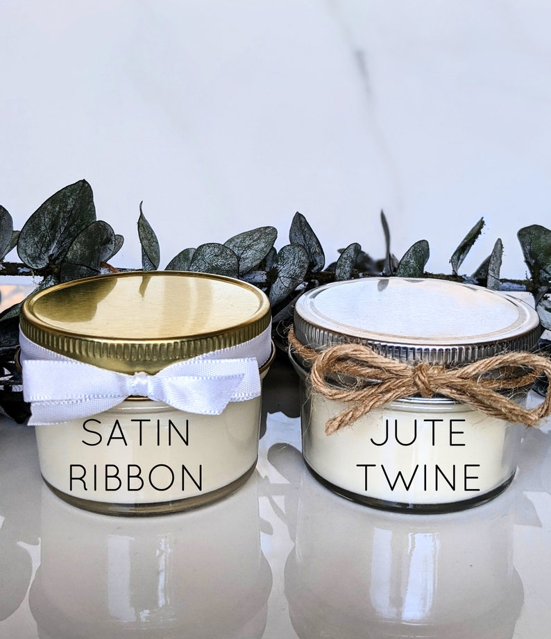 May include: Two small glass jars with white candles inside. The jars have gold and silver lids. The jar on the left has a white satin ribbon tied around the lid. The jar on the right has a brown jute twine tied around the lid. The jars are labeled "SATIN RIBBON" and "JUTE TWINE".