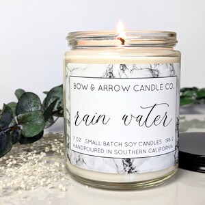 Op de afbeelding: Een glazen kaars pot met een wit marmerpatroon label. Het label luidt "BOW & ARROW CANDLE CO. rain water 7OZ SMALL BATCH SOY CANDLES 198 G HANDPOURED IN SOUTHERN CALIFORNIA".