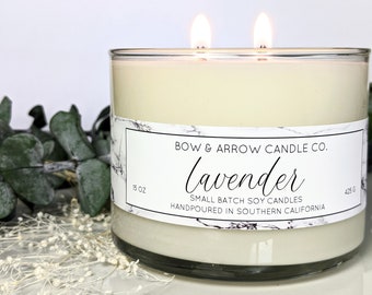 May include: A clear glass jar candle with a white label that reads "BOW & ARROW CANDLE CO. Lavender SMALL BATCH SOY CANDLES HANDPOURED IN SOUTHERN CALIFORNIA" with the text "15 OZ" and "425 G" on either side. The candle is lit with two wicks.