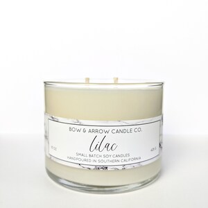 May include: A clear glass candle jar with a white soy candle inside. The label reads "BOW & ARROW CANDLE CO. Lilac SMALL BATCH SOY CANDLES HANDPOURED IN SOUTHERN CALIFORNIA".