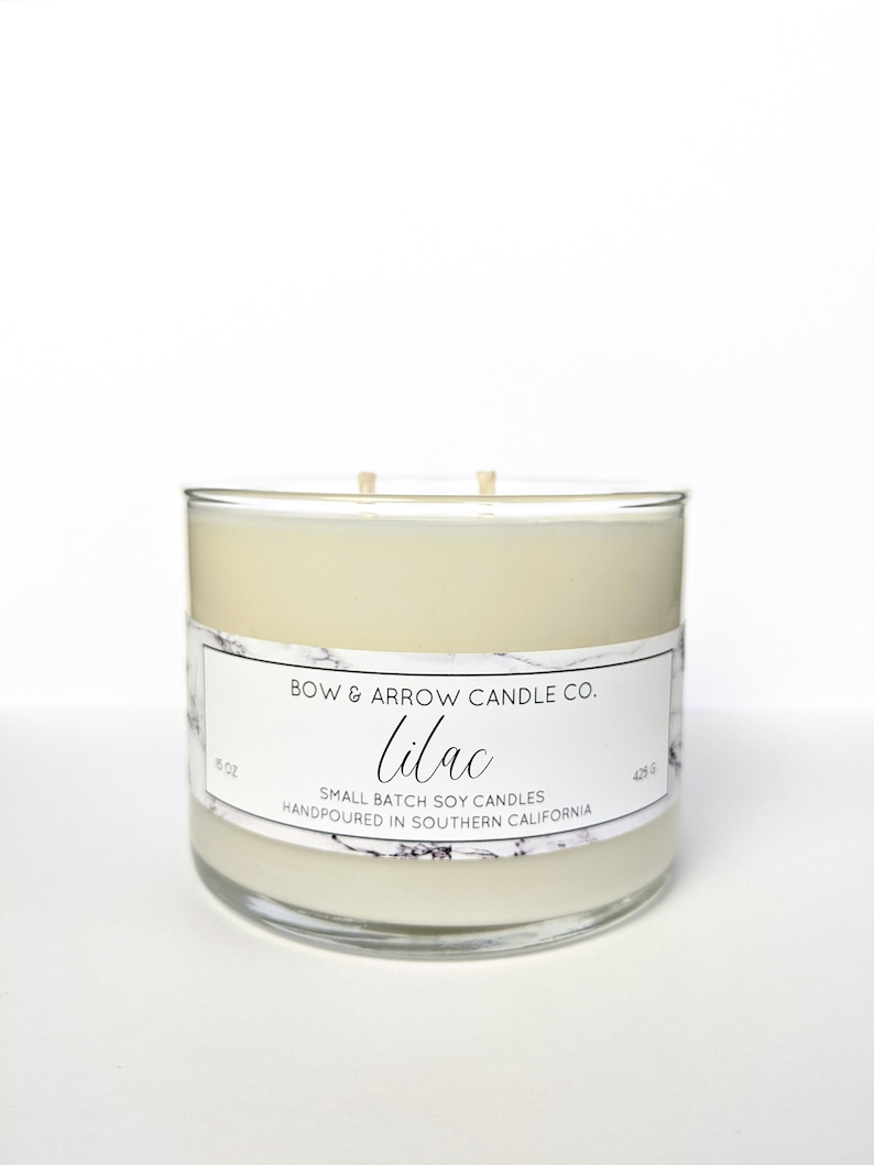 May include: A clear glass candle jar with a white soy candle inside. The label reads "BOW & ARROW CANDLE CO. Lilac SMALL BATCH SOY CANDLES HANDPOURED IN SOUTHERN CALIFORNIA".