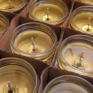 May include: Close-up of multiple glass jars filled with a golden yellow wax, each with a single wick centered in the jar. The jars are arranged in rows on a brown cardboard surface.