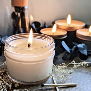 May include: A lit candle in a clear glass jar with a white wax base, surrounded by dried flowers and matches. There are four additional candles in the background, two of which are lit.