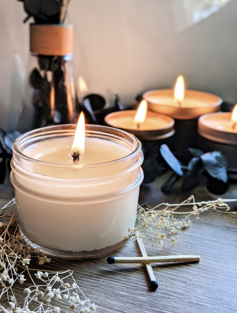 May include: A lit candle in a clear glass jar with a white wax base, surrounded by dried flowers and matches. There are four additional candles in the background, two of which are lit.