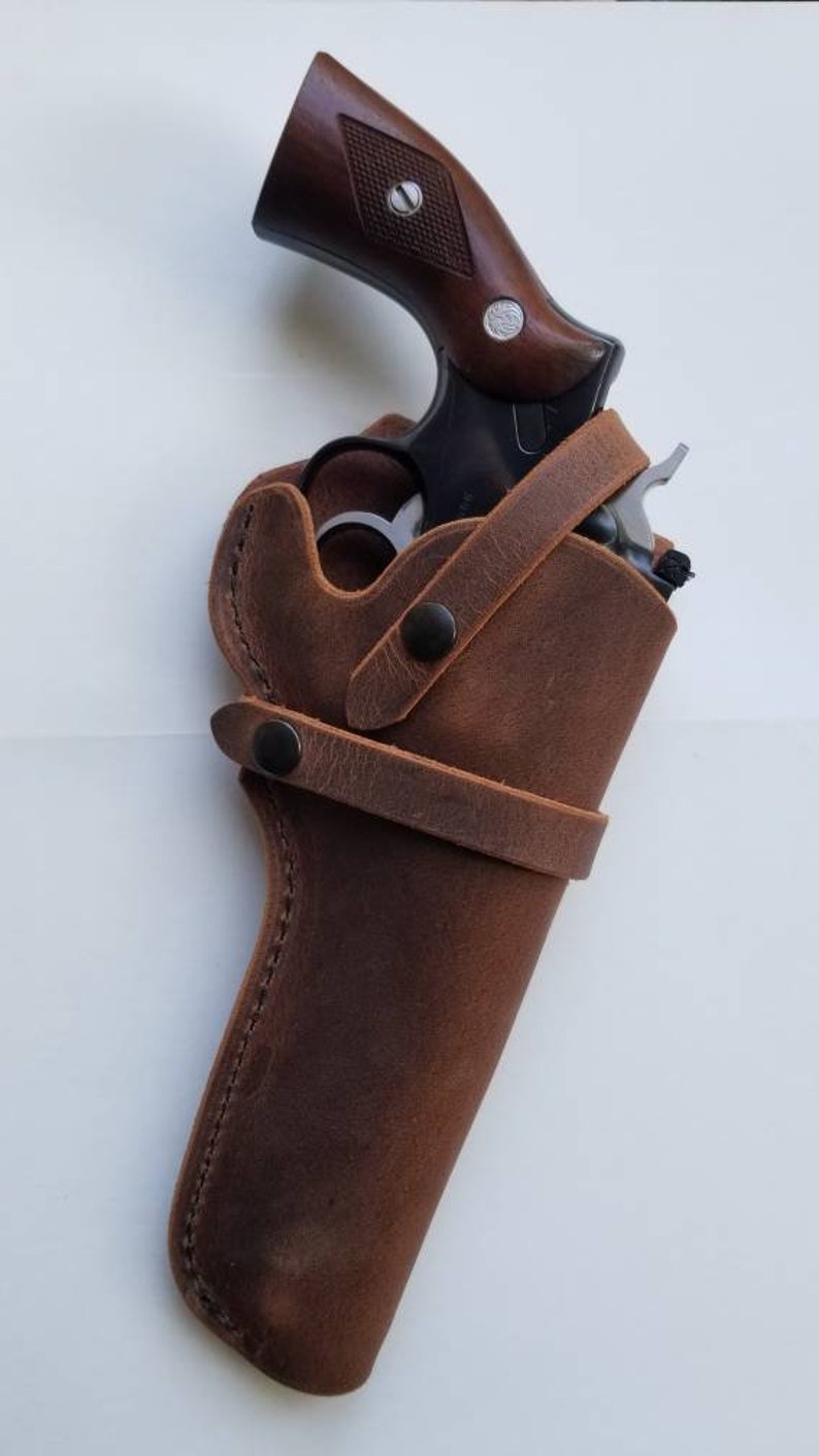 May include: A brown leather holster with a strap and snap closures. A black revolver is inside the holster.