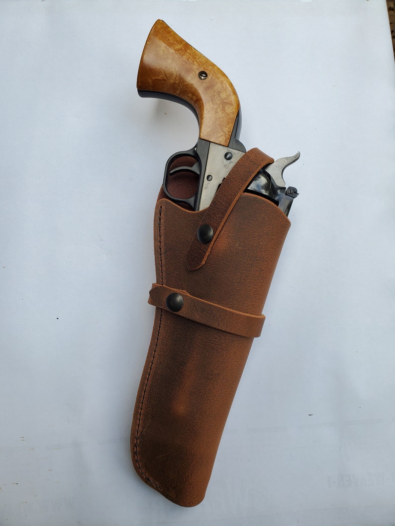 May include: A brown leather holster with a black revolver inside. The holster has a strap with a snap closure.