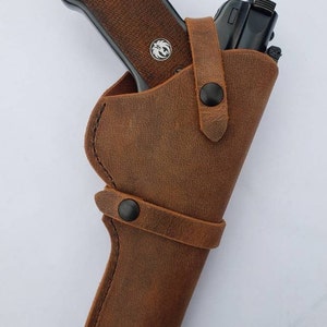 Puede incluir: Una funda de cuero marr&oacute;n con una pistola negra. La funda tiene dos correas con broches negros. La pistola tiene una empu&ntilde;adura marr&oacute;n y un emblema plateado. La funda est&aacute; dise&ntilde;ada para llevar un arma de fuego.