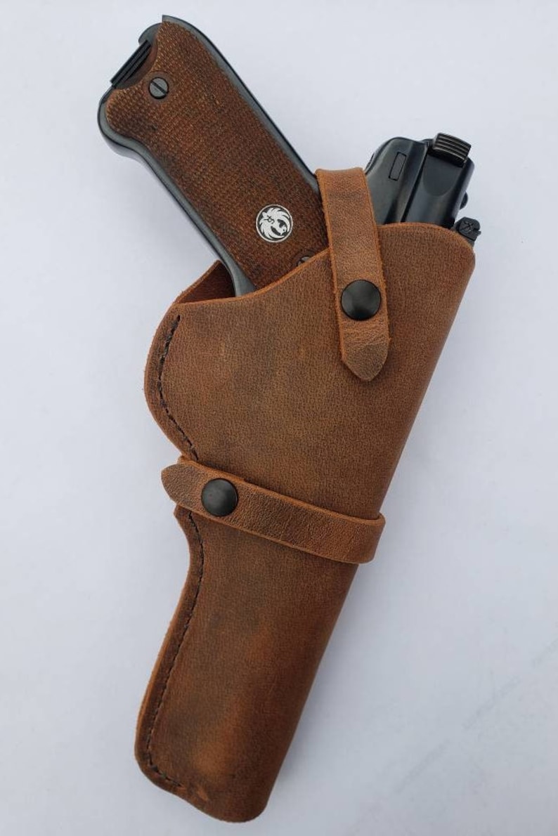Puede incluir: Una funda de cuero marr&oacute;n con una pistola negra. La funda tiene dos correas con broches negros. La pistola tiene una empu&ntilde;adura marr&oacute;n y un emblema plateado. La funda est&aacute; dise&ntilde;ada para llevar un arma de fuego.
