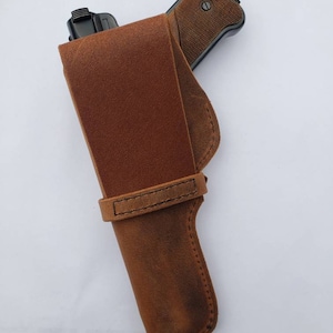Puede incluir: Una funda de cuero marr&oacute;n con un dise&ntilde;o cosido, que sostiene una pistola negra. La funda tiene una trabilla para el cintur&oacute;n y una solapa para asegurar el arma de fuego. La empu&ntilde;adura del arma es visible, con un patr&oacute;n texturizado.