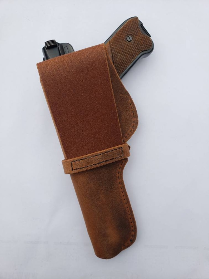 Puede incluir: Una funda de cuero marr&oacute;n con un dise&ntilde;o cosido, que sostiene una pistola negra. La funda tiene una trabilla para el cintur&oacute;n y una solapa para asegurar el arma de fuego. La empu&ntilde;adura del arma es visible, con un patr&oacute;n texturizado.