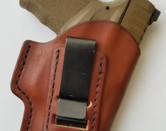 May include: A brown leather holster with a black metal clip for a handgun. The holster has a stitched design and a brown leather strap.