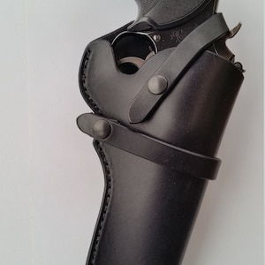 May include: A black revolver handgun with a textured grip is partially inserted into a black leather holster. The holster has a belt loop and a strap with a snap closure. The gun has a gold emblem on the grip.