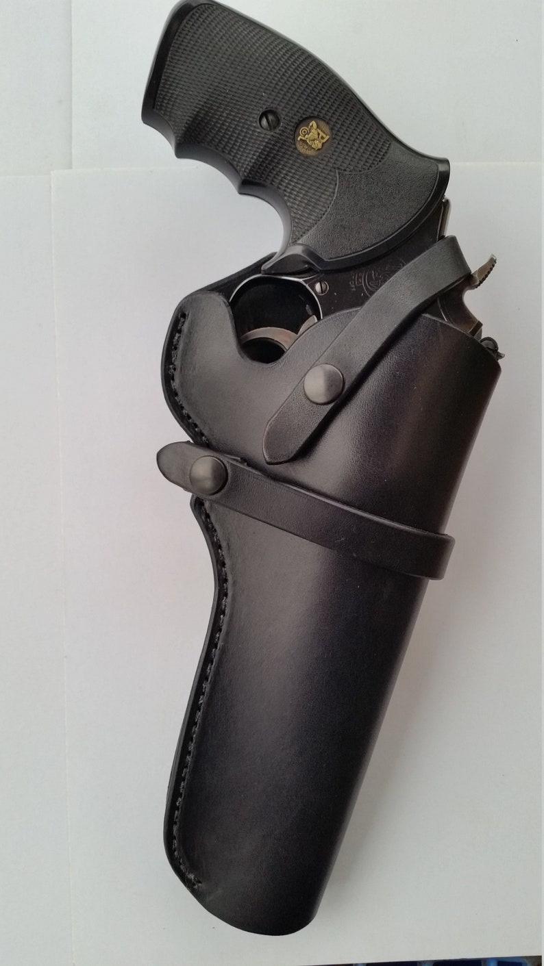May include: A black revolver handgun with a textured grip is partially inserted into a black leather holster. The holster has a belt loop and a strap with a snap closure. The gun has a gold emblem on the grip.