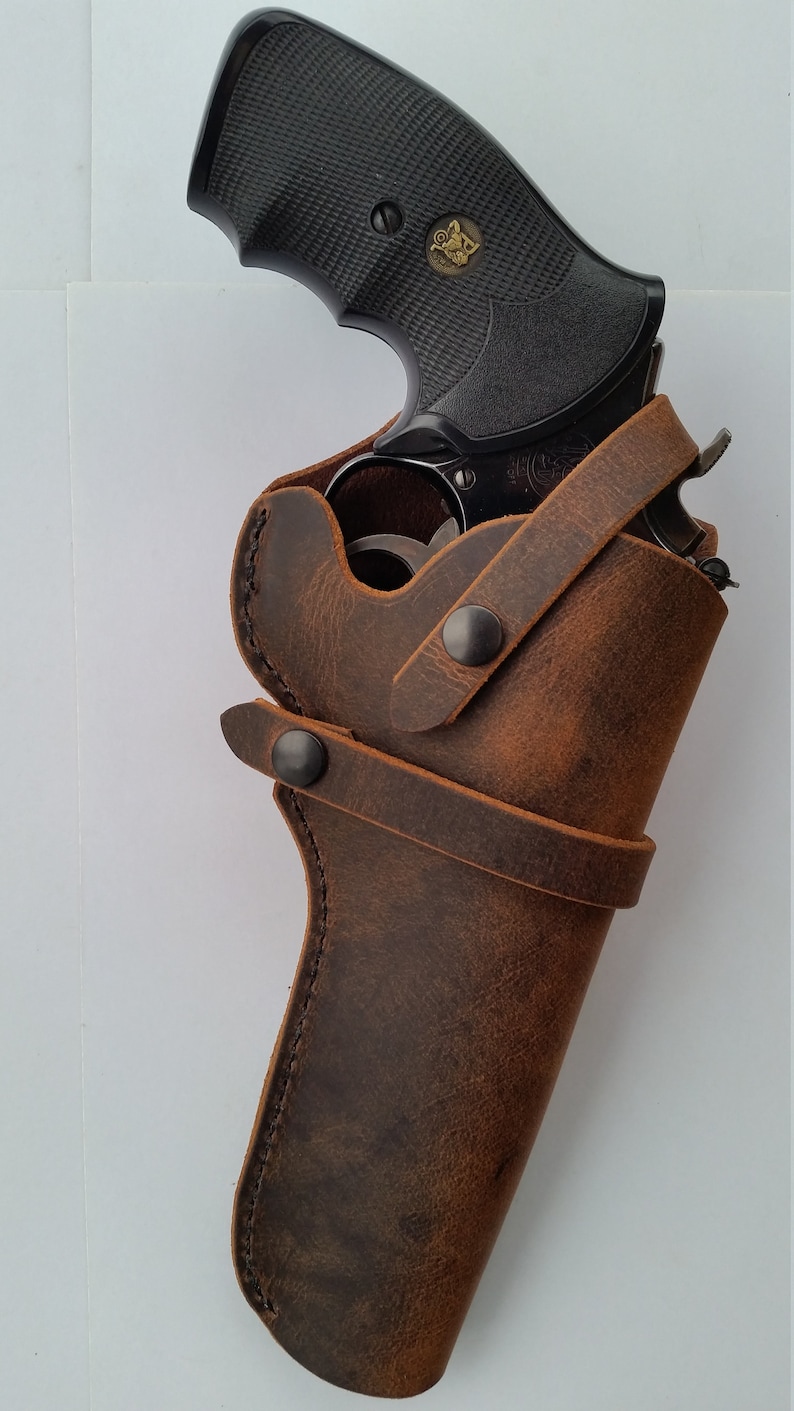 May include: A brown leather holster with a black revolver inside. The holster has a strap with two metal snaps.
