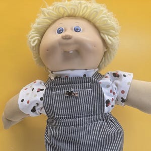 May include: A doll with light skin, blue eyes, and curly blonde yarn hair. It wears a white short-sleeved shirt featuring a pattern of small red and black polka dots and mouse silhouettes, paired with dark gray pinstriped denim overalls with a metal pin.