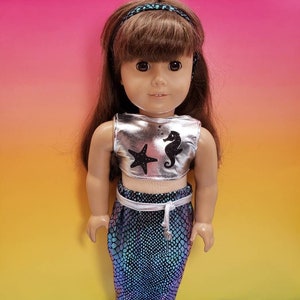 May include: A silver mermaid costume for an 18-inch doll. The costume includes a silver top with a black star and seahorse design, a blue and green iridescent mermaid tail, and silver fin feet.