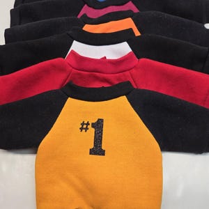 May include: A stack of yellow, red, white, orange, maroon, and blue sweatshirts with black sleeves and a black waistband. The bottom sweatshirt has a black number "1" with glitter on the back.