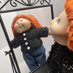 May include: A Cabbage Patch Kid doll with orange hair and blue eyes, wearing a black sweater with white buttons and blue jeans, looking into a black framed mirror. The mirror has a decorative top.