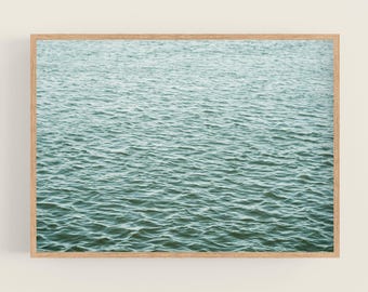 May include: A rectangular wall art print featuring a high-angle view of rippling water in shades of teal and deep green, enclosed within a light-toned wooden frame that borders the entire image with a clean, simple edge.