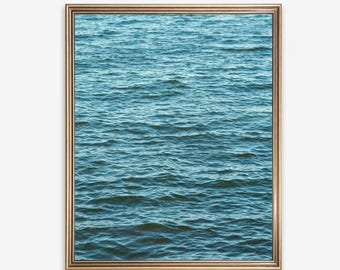 Op de afbeelding: Een rechthoekig wandkunstwerk met een foto van kabbelend blauw water. De afbeelding is ingelijst in een dunne, goudkleurige metalen lijst met een eenvoudig profiel, die het getextureerde oppervlak van het water in diepblauwe en groenblauwe tinten toont.