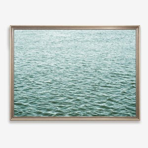 May include: A rectangular wall art piece featuring a photograph of rippling water in shades of teal, blue, and green. The image is enclosed within a thin, metallic-toned frame that provides a clean border around the textured aquatic scene.