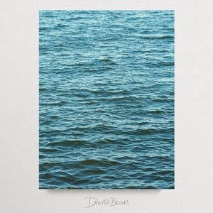Op de afbeelding: Een rechthoekige print met een close-up van kabbelend water in tinten blauw en blauwgroen. Het wateroppervlak is getextureerd met kleine golven, waardoor een gevoel van diepte en beweging ontstaat. De handtekening van de kunstenaar, "DANITA BROOKS", staat onderaan.