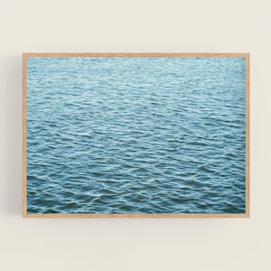May include: A framed print featuring a close-up view of rippling blue water. The water's surface is textured with small waves, creating a sense of depth and movement. The wooden frame adds a natural touch to the artwork.